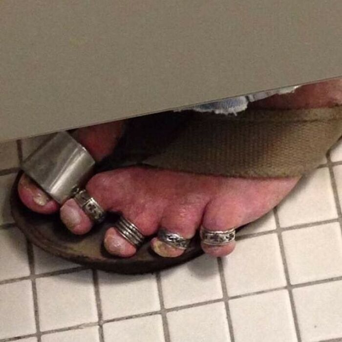 Bizarre photo of feet wearing rings and a sandal in a restroom stall setting.