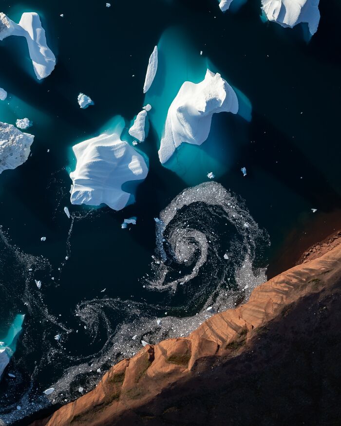 Aerial view of icy waters with swirling patterns and icebergs near a rugged coastline, showcasing untamed nature.