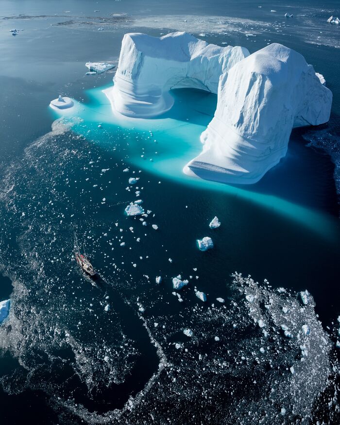 Aerial view of a large iceberg with a sailboat navigating icy waters, highlighting the untamed beauty of nature.