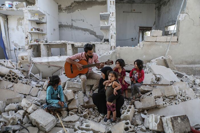 Man playing guitar for children in rubble, showcasing resilience, Tokyo International Foto Awards.