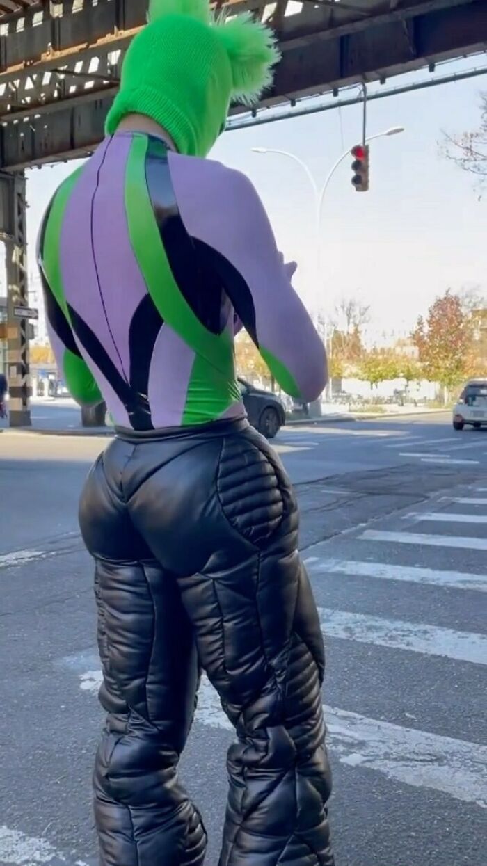 NYC local in bold fashion: green hood, purple and green top, quilted black pants on city street.