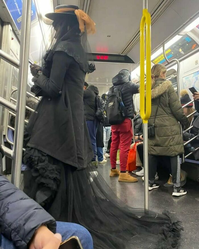 New Yorker in a long black dress and hat with veil, riding the subway, showcasing experimental fashion.