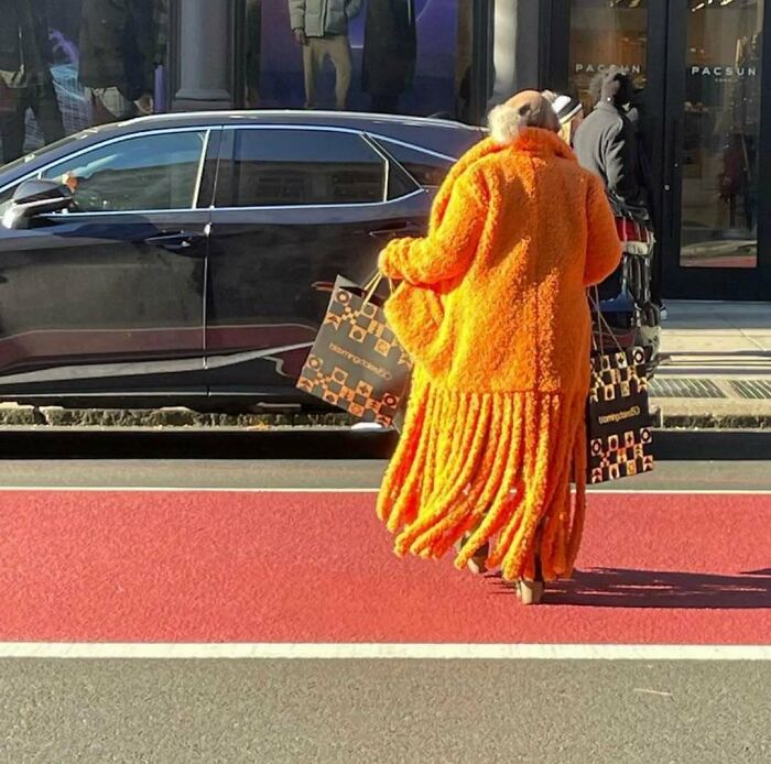 NYC local in a bold orange coat holds shopping bags, showcasing experimental fashion on a city street.