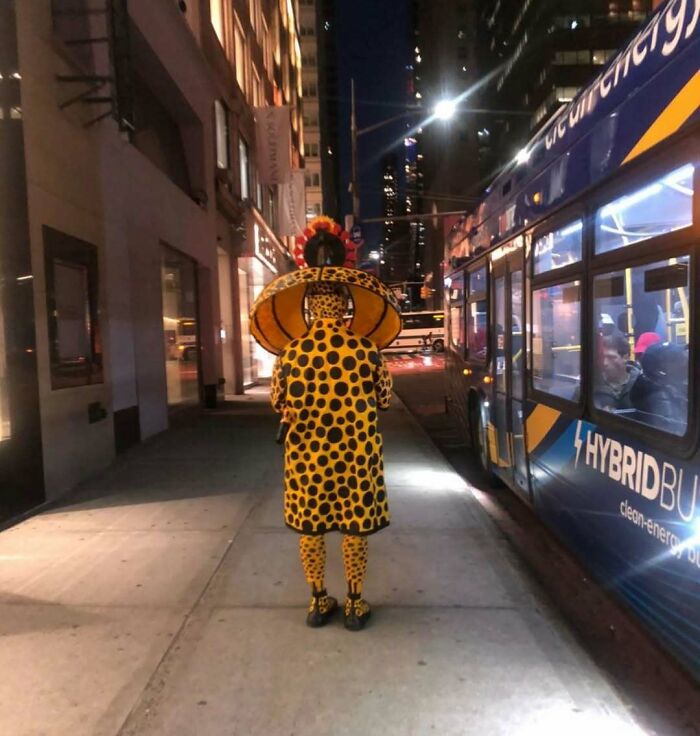 NYC local in a bold polka dot outfit with large hat on a city sidewalk at night, experimenting with unique fashion.