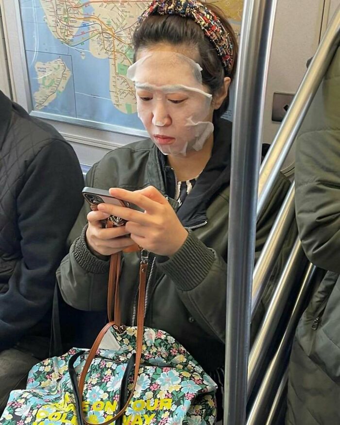 Passenger on subway with face mask, using phone and carrying floral bag; amusing scene in public transit.