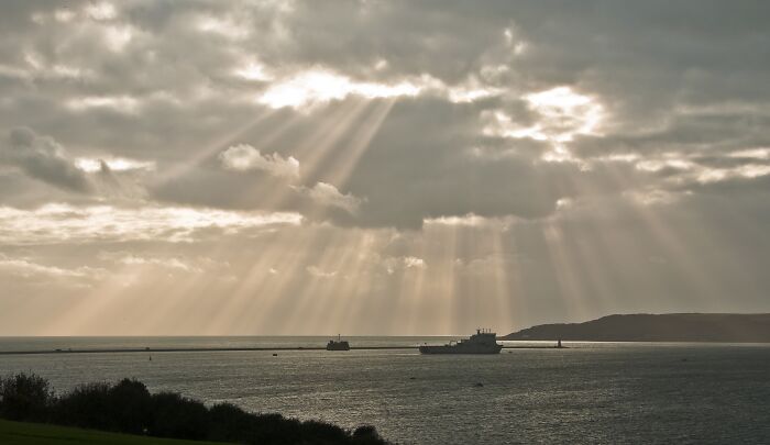 Sunbeams breaking through clouds over the ocean, highlighting natural phenomena with boats in the distance.