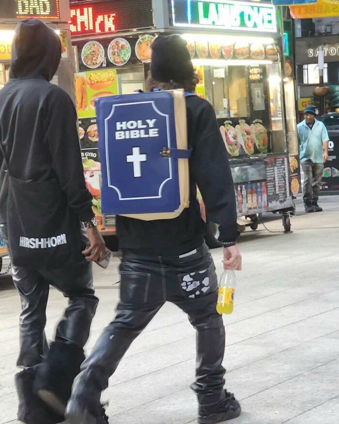 NYC local wearing a bold Holy Bible backpack, walking in front of a street food cart, showcasing experimental fashion.