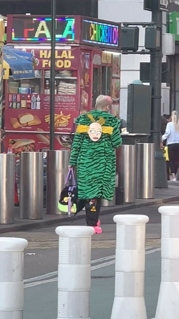 NYC local wearing a bold green patterned coat with unique face design, holding colorful bags near a street food cart.