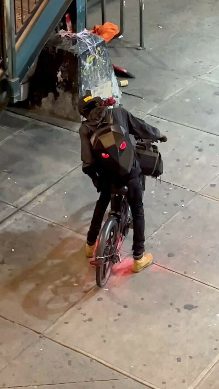 NYC local in experimental fashion on a bicycle at night, wearing a black outfit with glowing red backpack eyes.