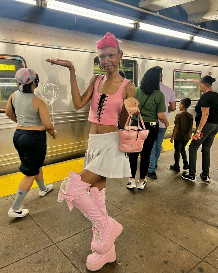 NYC local on subway platform wearing pink top, skirt, and boots, showcasing experimental fashion style.