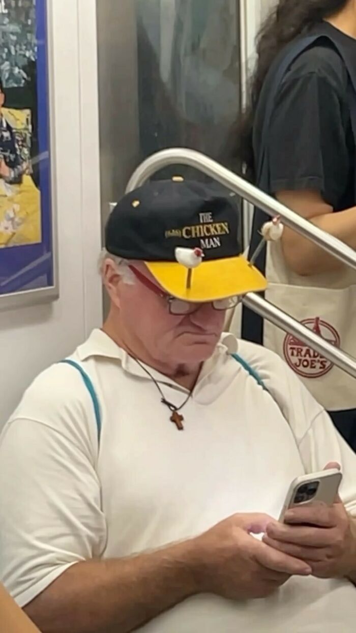 NYC local on the subway, wearing a unique "Chicken Man" hat while using a phone, showcasing New York fashion experimentation.