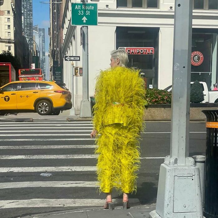 NYC local in bold, yellow feathered outfit stands on a city street, showcasing experimental New York fashion.