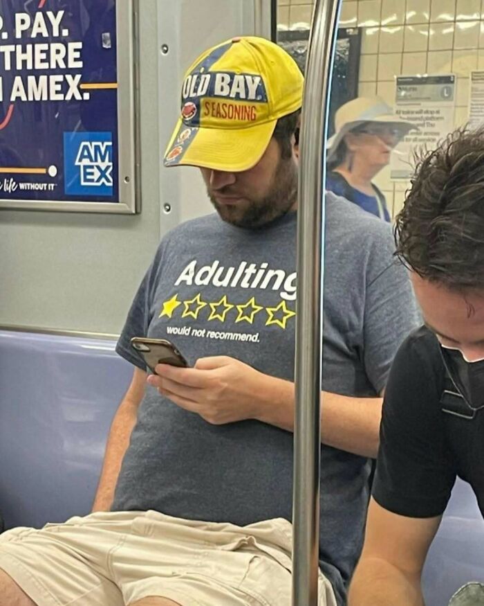 Subway passenger wearing "Adulting" shirt and funny hat, looking at phone.