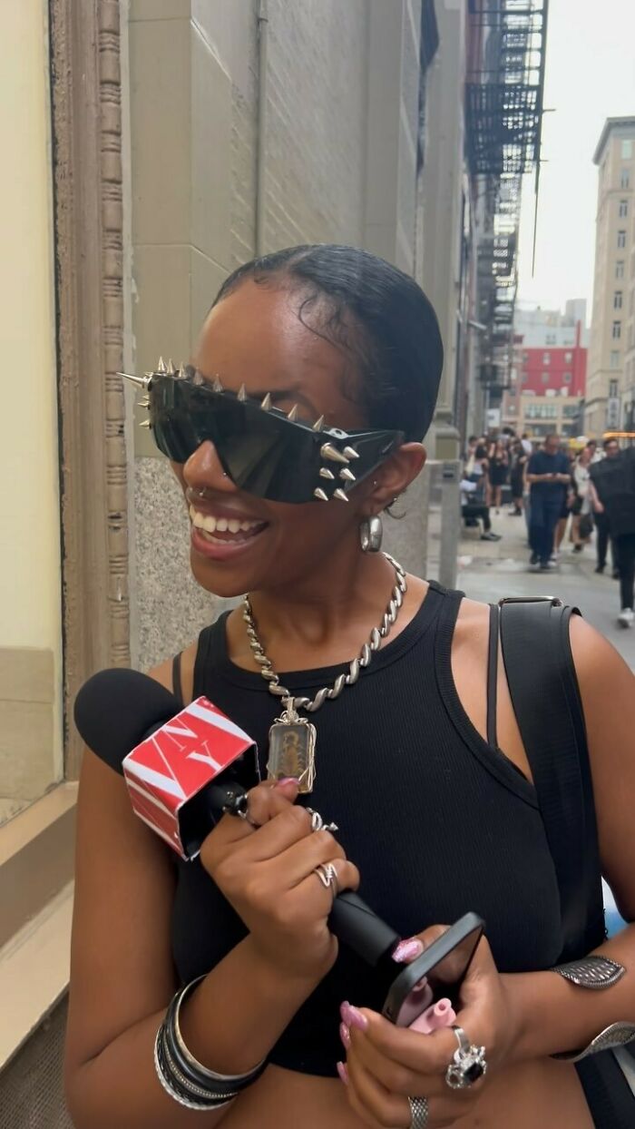 NYC local in spiked sunglasses and black top, holding a microphone, embracing experimental fashion on the street.