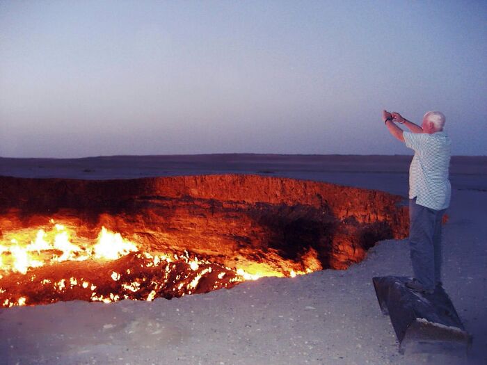 Person photographing the blazing Darvaza gas crater at dusk, showcasing strange natural phenomena.