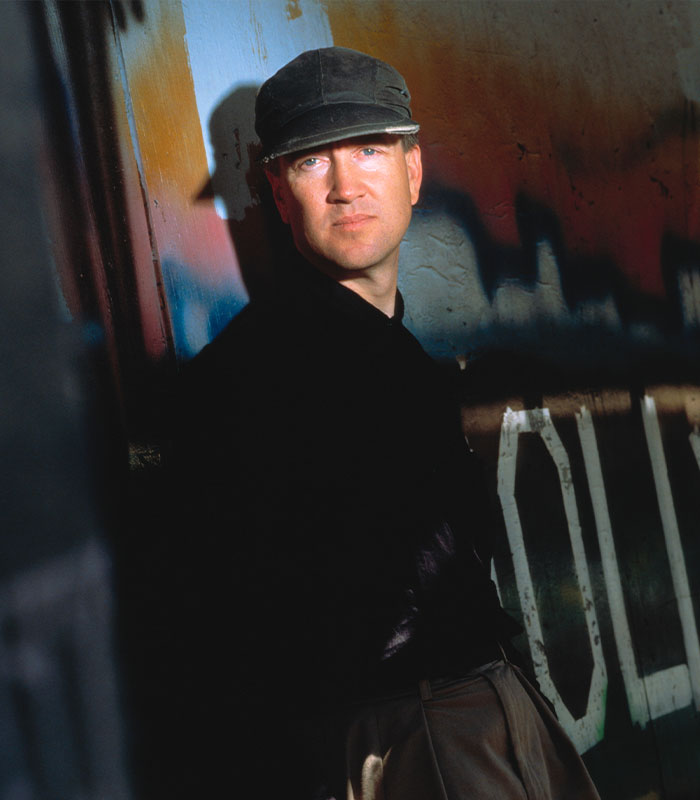 "Man in a cap and dark attire standing by a colorful graffiti wall, representing David Lynch quotes and reality themes."