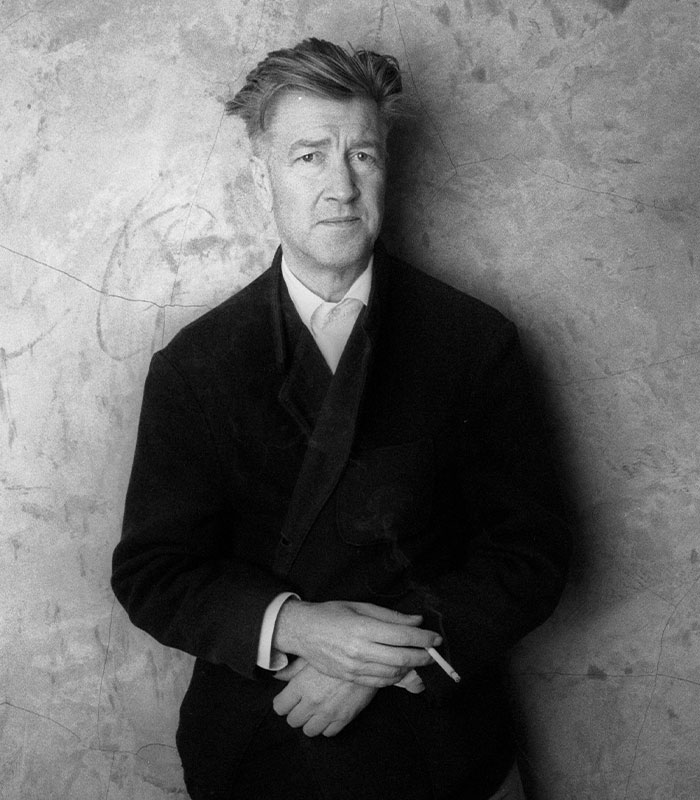 David Lynch holding a cigarette, dressed in a dark jacket, with a textured wall in the background.