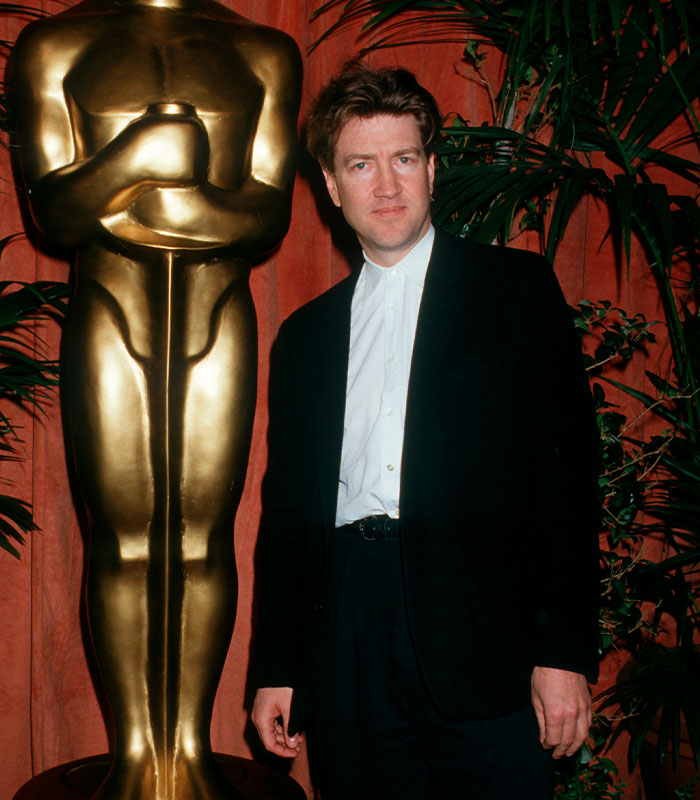 Man in a suit standing next to a large gold statue, surrounded by plants, representing David Lynch quotes theme.