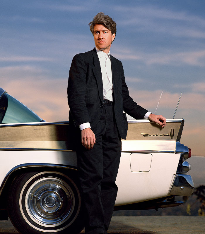 David Lynch standing beside a classic car, wearing a black suit, with a serene sunset background.