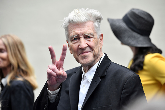 A man in a suit with white hair gestures a peace sign, surrounded by people, celebrating David Lynch's work.