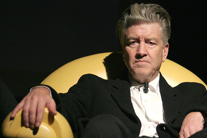 Director David Lynch seated in a yellow chair, wearing a black jacket and white shirt, against a dark background.