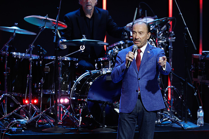 Performer singing at inauguration with drummer in the background, wearing a blue blazer and red tie.