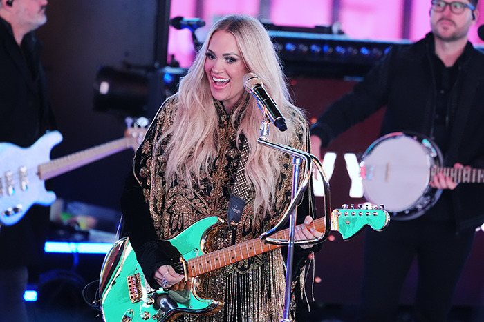 Singer performing on stage with a guitar, wearing an embellished outfit, during a big inauguration event.