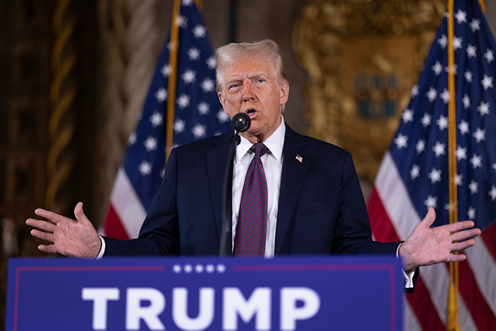 A man speaking at a podium with "TRUMP" sign in front, against a backdrop of American flags.