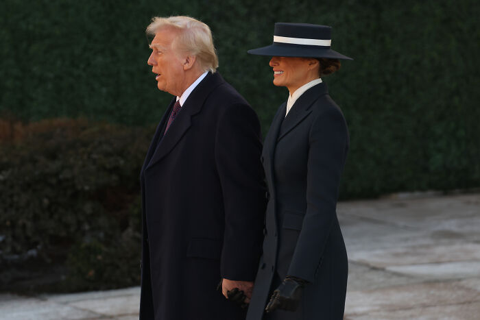 A woman in a dark coat and hat walks beside a man in a black overcoat, stirring body double rumors.