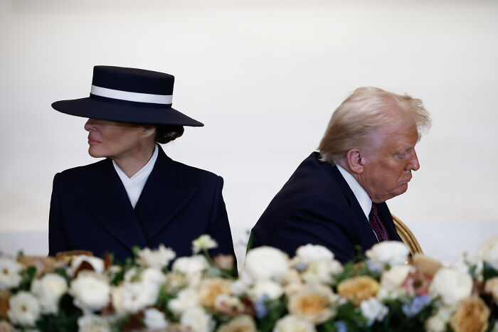 Two individuals sit back-to-back surrounded by flowers, with one wearing a dark hat and coat.