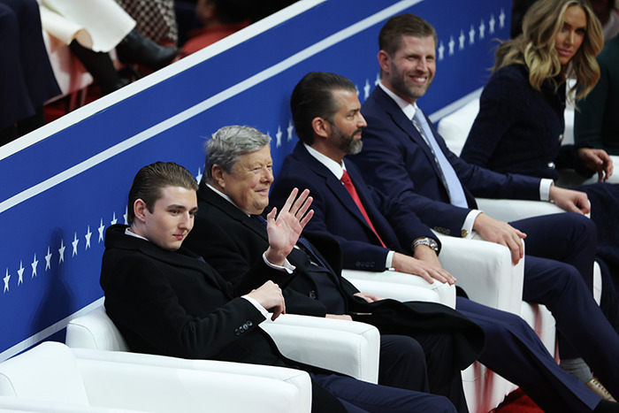 Barron Trump sits on a white chair at the inauguration, waving.