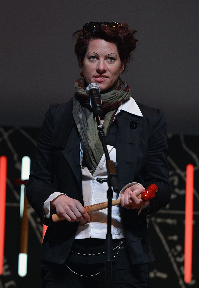 A woman in a black jacket and scarf holding a microphone on stage, related to Neil Gaiman's ex-wife.