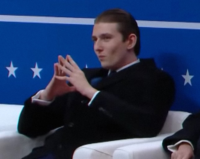 Barron Trump in a suit making a significant hand gesture during an inaugural event.