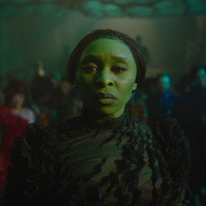 Cynthia Erivo as Elphaba standing in a dimly lit crowd, wearing a textured dark outfit.