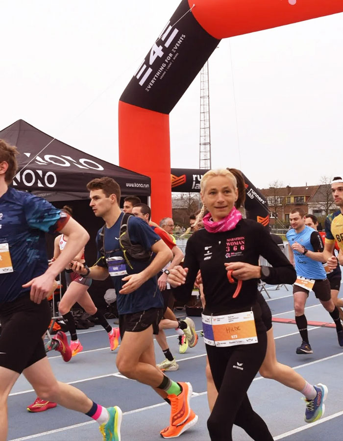 55YO woman running marathon, setting world record for 366 marathons in a year, wearing race bib and pink scarf.