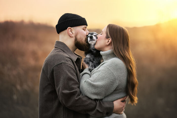 A couple kisses their dog in a sunset landscape, celebrating pet photography award.