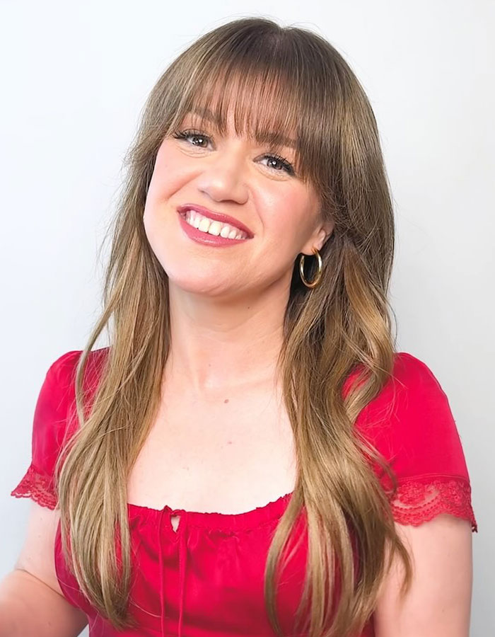 Person in a red top smiling, with long hair and hoop earrings, related to Hollywood beauty trends.