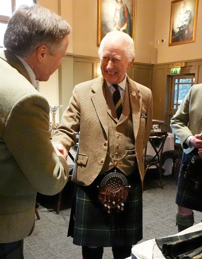 Man in a tweed jacket and kilt shakes hands indoors, a portrait of King Charles hangs in the background.