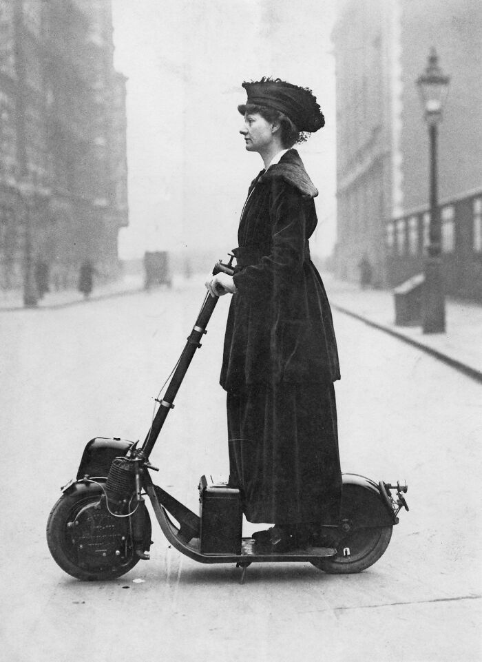 A woman in vintage attire riding an early motorized scooter, showcasing historical inventions.