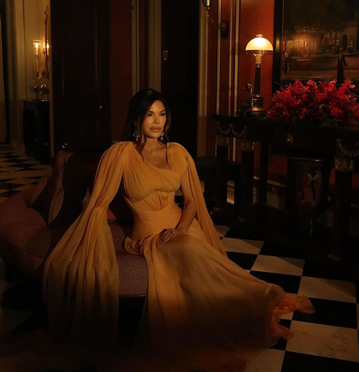 Lauren Sanchez in an elegant setting, wearing a flowing gown, seated on a sofa with ambient lighting.