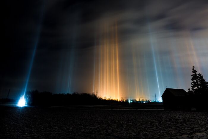 Pillars of light in the night sky, a stunning natural phenomenon over a rural landscape with a barn and trees.