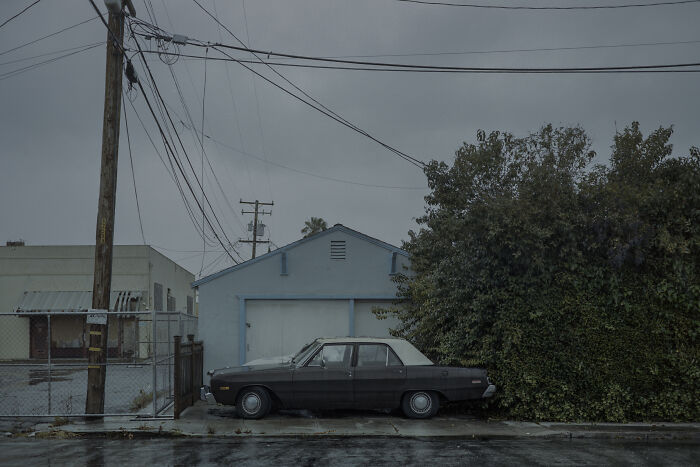 Suburban disconnection: an old car parked in front of a small garage on a rainy day, surrounded by overgrown bushes.