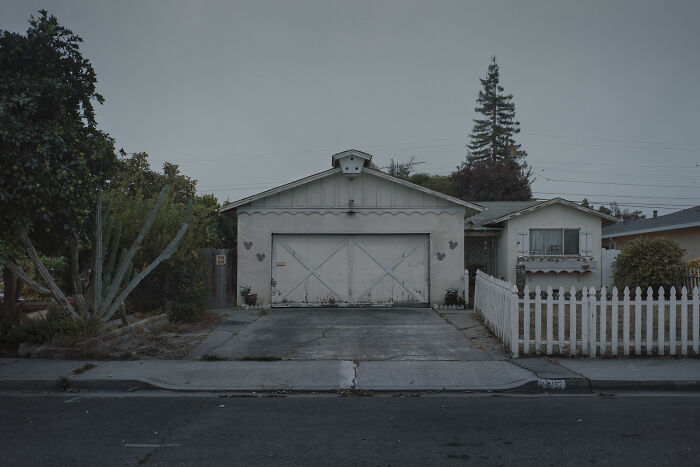 Suburban disconnection captured in a quiet, fading neighborhood scene with a white picket fence and a worn-out garage.