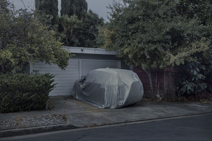 Covered car on a suburban driveway, surrounded by trees, capturing the essence of suburban disconnection.