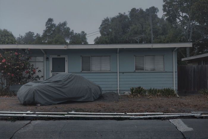 Suburban disconnection depicted by a covered car in front of a muted blue house, surrounded by trees under a gray sky.