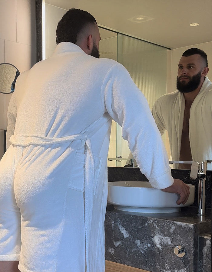 Man in a white robe looks at his reflection in a bathroom mirror, related to male stripper experiences at bachelorette parties.