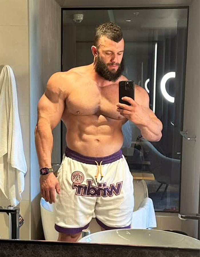 Muscular man in shorts taking a mirror selfie in a well-lit room before an event.