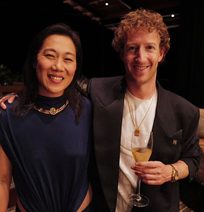 Man and woman smiling at a formal event, man holding a wine glass, both wearing elegant attire. Mark Zuckerberg terrain.