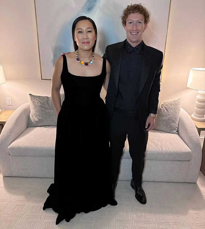 Man in formal attire next to a woman in a black dress, both smiling in a softly lit room.