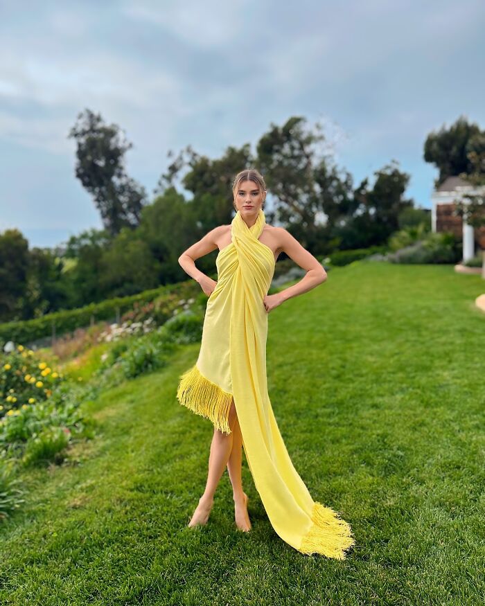 Fashion prodigy model in a flowing yellow dress poses gracefully on a lush green lawn.